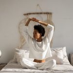 A woman sitting cross-legged and stretching in a tranquil bedroom setting, capturing a peaceful morning moment.