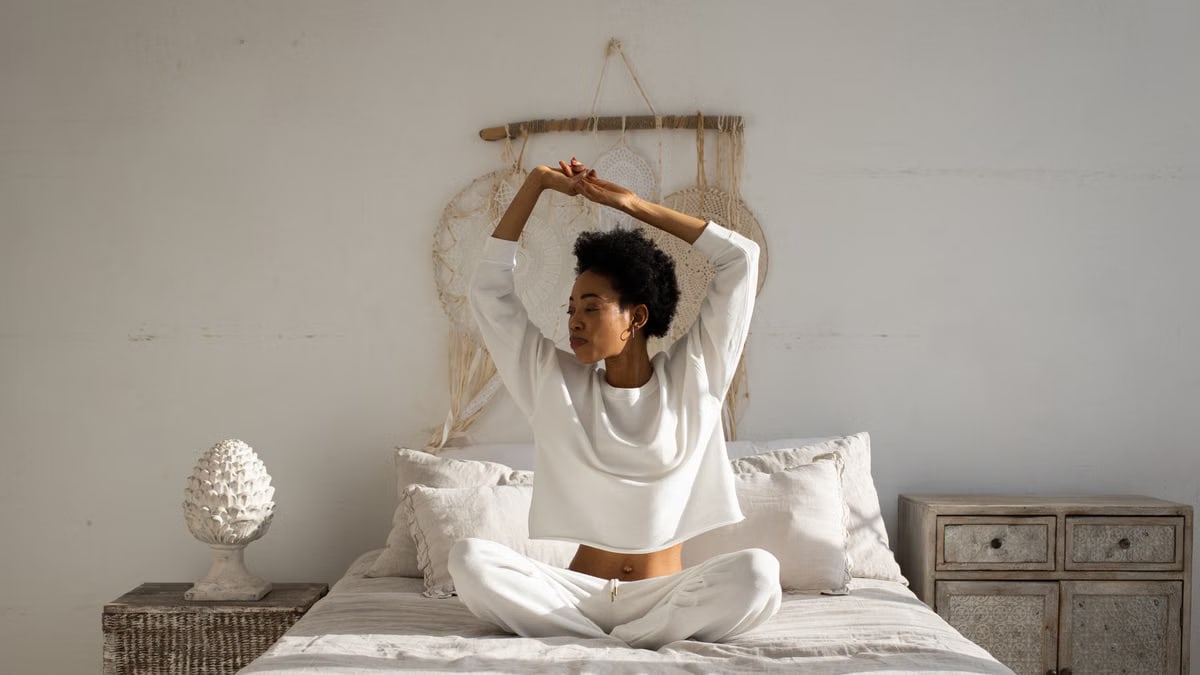 A woman sitting cross-legged and stretching in a tranquil bedroom setting, capturing a peaceful morning moment.