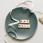 Flat lay of scrabble tiles spelling 'eat well' on a plate with a tape measure, promoting healthy eating.
