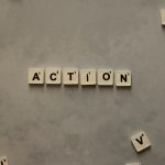 Flat lay of Scrabble tiles spelling 'Action' on a gray background, surrounded by random letters.